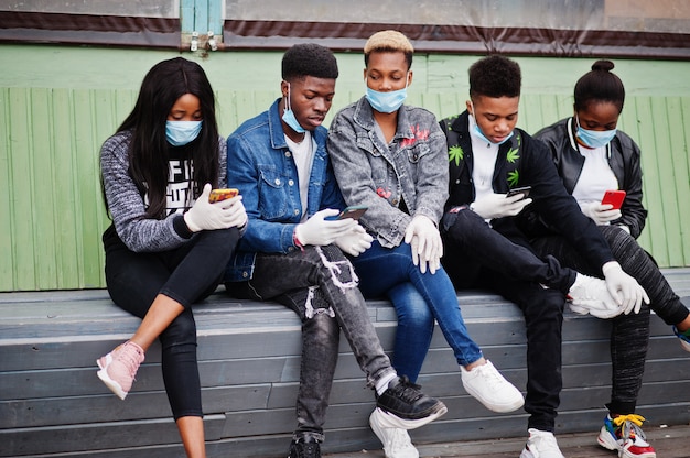 Grupo de amigos adolescentes africanos sentado com telefones, usando máscaras médicas proteger de quarentena de vírus de coronavírus infecções e doenças.