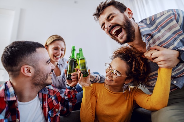 Grupo de amigos alegres se divertindo em casa, brindando com cerveja e rindo.