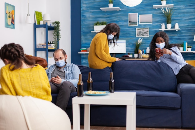 Grupo de amigos anti-sociais multiétnicos mantendo o distanciamento social navegando em smartphones na sala de estar de um apartamento durante a pandemia global de coronavírus.