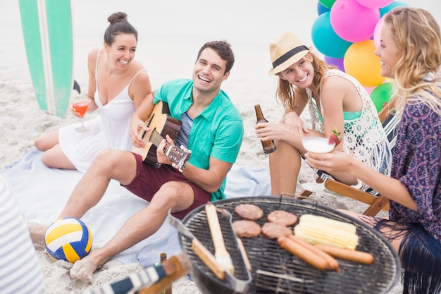 Grupo de amigos com bebidas e violão, sentado ao lado de um churrasco