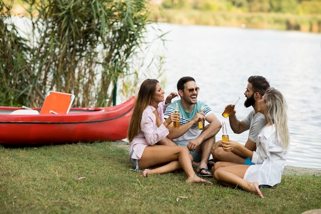 Grupo de amigos com garrafas de cidra sentado perto do barco perto do belo lago e se divertindo