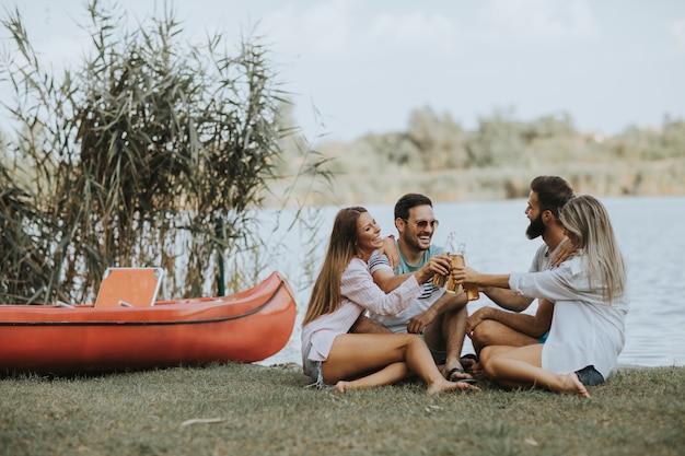 Grupo de amigos com garrafas de cidra sentado perto do barco perto do belo lago e se divertindo