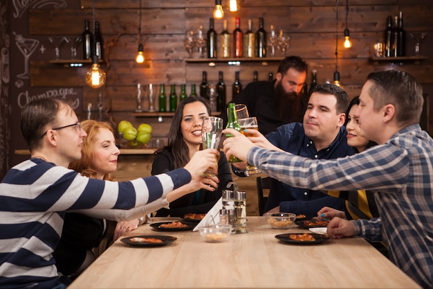 Grupo de amigos felizes bebendo cerveja e comendo pizza no bar restaurante. Eles estão sentados em uma mesa de madeira.