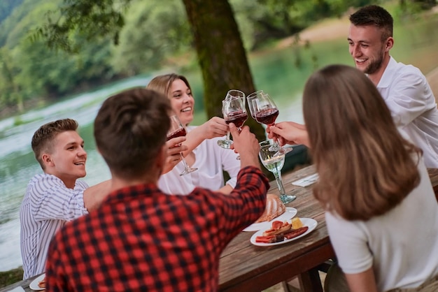 grupo de amigos felizes brindando com copo de vinho tinto enquanto fazia piquenique jantar francês ao ar livre durante as férias de verão perto do rio na bela natureza