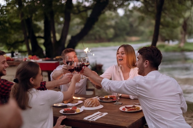 grupo de amigos felizes brindando com copo de vinho tinto enquanto fazia piquenique jantar francês ao ar livre durante as férias de verão perto do rio na bela natureza