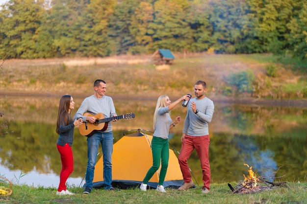 Grupo de amigos felizes com guitarra, se divertindo ao ar livre, dançando e pulando perto do lago no fundo do parque, o lindo céu. diversão em camping