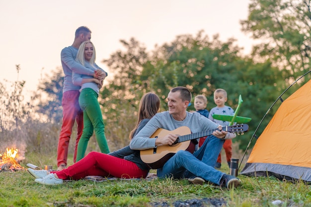 Grupo de amigos felizes com guitarra, se divertindo ao ar livre, perto da barraca da fogueira e do turista. Diversão de acampamento família feliz