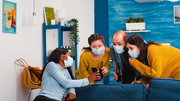 Grupo de amigos multiétnicos na sala de estar assistindo a um clipe no smartphone, sentado no sofá, respeitando o distanciamento social, usando máscara facial durante a pandemia global na nova festa normal