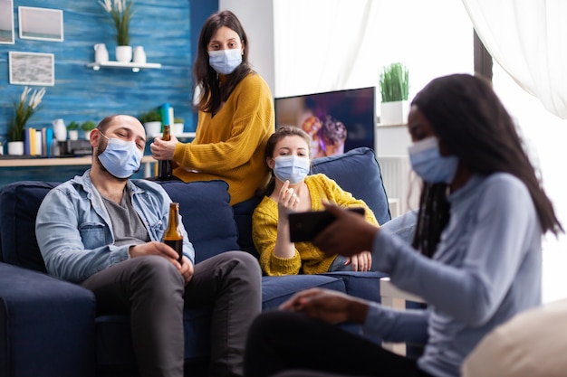 Grupo de amigos multiétnicos na sala de estar assistindo clipe no smartphone, sentado no sofá, respeitando o distanciamento social, usando máscara facial durante a pandemia global.