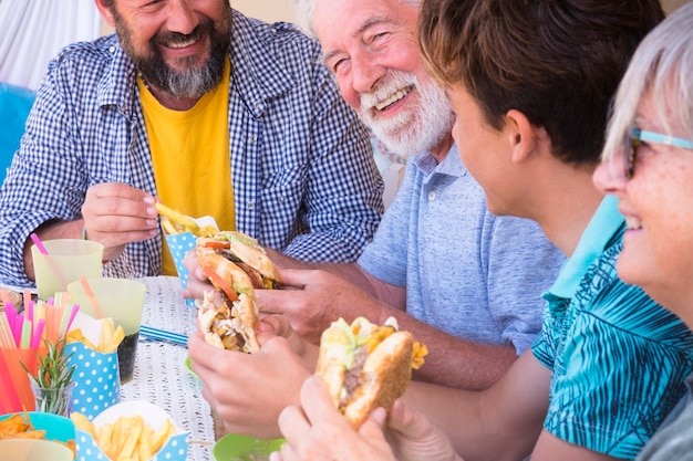 Grupo de amigos ou família almoçando juntos com hambúrguer e batatas fritas juntos - pessoas felizes celebrando juntos - com lixo e comida não saudável - gerações mistas