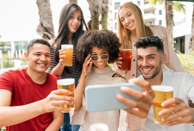 Grupo de amigos posando para uma selfie com canecas de cerveja gelada erguendo os copos para um brinde para a câmera no celular com sorrisos felizes