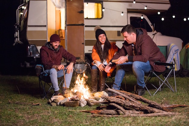 Grupo de amigos relaxando e rindo juntos ao redor da fogueira nas montanhas. Carrinha de campista retro.
