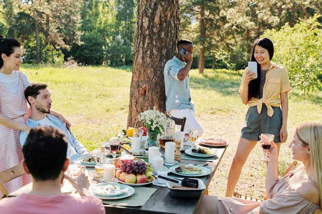 Foto grupo de amigos repousantes relaxando na mesa servida após o jantar ao ar livre sob um pinheiro enquanto uma garota asiática tira fotos deles no smartphone