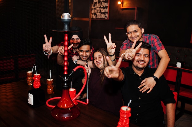 Grupo de amigos se divertindo e descansar na boate, beber cocktails e fumar narguilé mãos com dois dedos para cima.