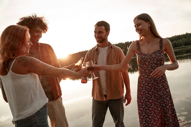 Grupo de amigos se divertindo em um piquenique perto de um lago ou rio, bebendo vinho, cerveja, copos de cidra tilintando enquanto diz um brinde. Jovens a sorrir festas ao ar livre durante o pôr do sol na zona rural.