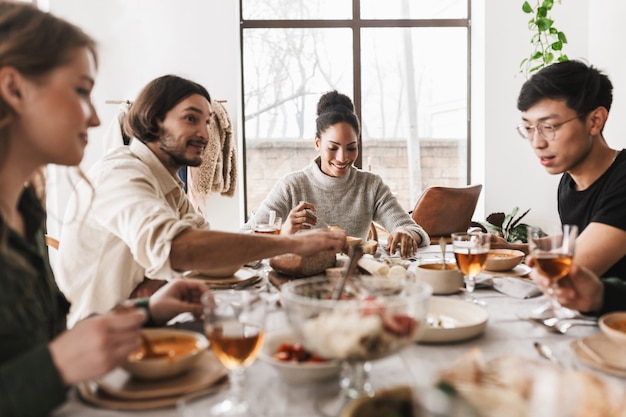 Grupo de atraentes amigos internacionais sentados à mesa cheia de comida e passando o tempo em um café aconchegante