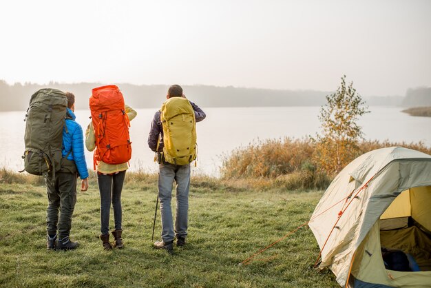 Grupo de caminhantes com mochilas coloridas em pé no lago perto do acampamento durante o nevoeiro