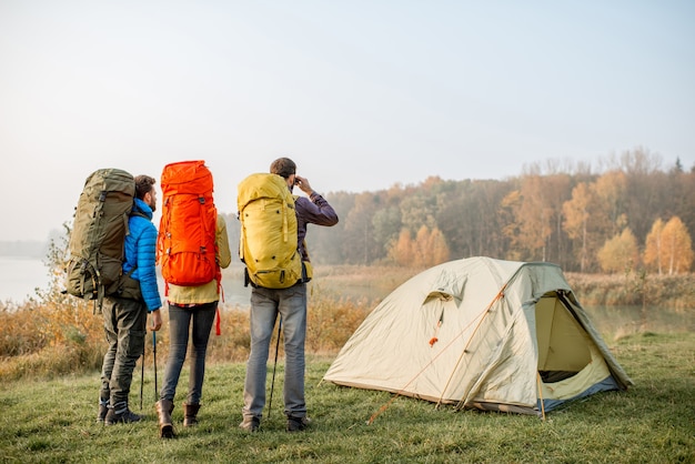 Grupo de caminhantes com mochilas coloridas em pé no lago perto do acampamento durante o nevoeiro