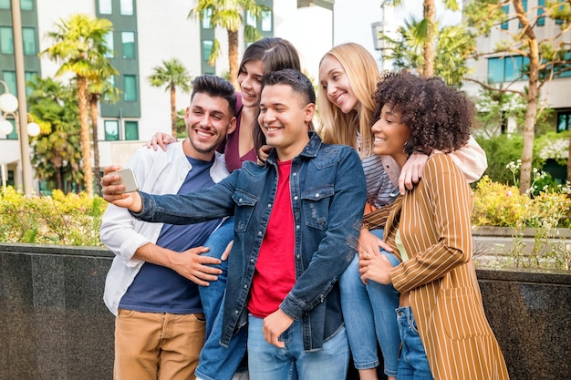 Grupo de cinco amigos multiculturais felizes tirando uma selfie em um telefone celular em uma rua da cidade rindo e sorrindo enquanto posam para a câmera