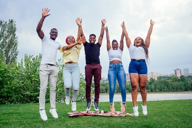 Grupo de cinco homens e mulheres afro-americanos sorridentes caminhando fora do tempo nublado perto do lago, estudantes de intercâmbio no trabalho em equipe da Rússia