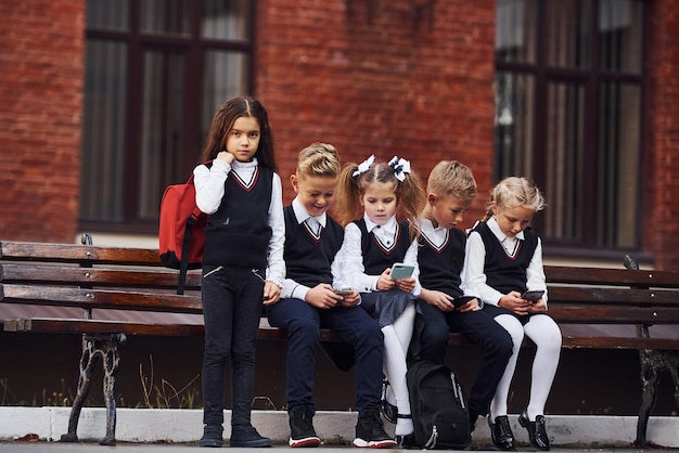 Grupo de crianças com uniforme escolar se senta no banco ao ar livre juntos, perto do prédio da educação.