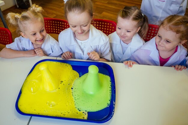 Grupo de crianças da escola com professor no laboratório da escola fazendo experimento observando a reação química com o corante com vinagre e vulcão de refrigerante usando vidro uniforme de vestido branco