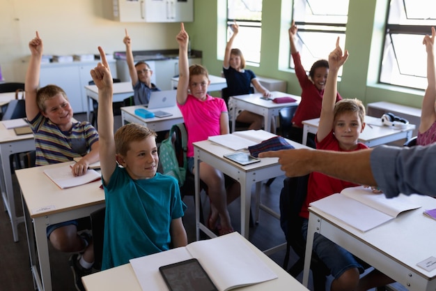 Grupo de crianças em idade escolar sentadas em carteiras e levantando as mãos para responder