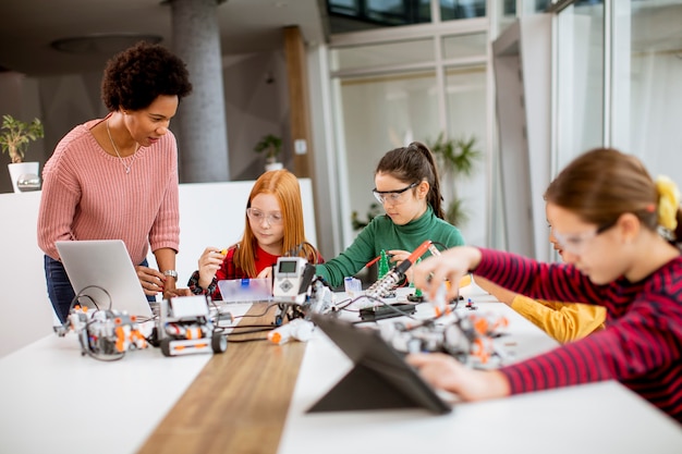 Grupo de crianças felizes com sua professora de ciências afro-americana com um laptop programando brinquedos elétricos e robôs na sala de aula de robótica