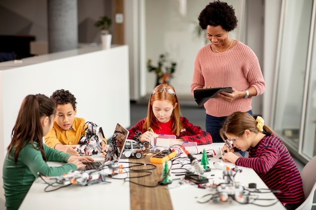 Grupo de crianças felizes com sua professora de ciências afro-americana com um laptop programando brinquedos elétricos e robôs na sala de aula de robótica