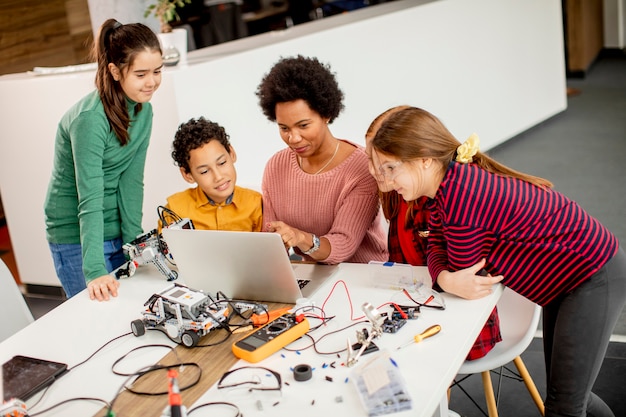 Foto grupo de crianças felizes com sua professora de ciências afro-americana com um laptop programando brinquedos elétricos e robôs na sala de aula de robótica