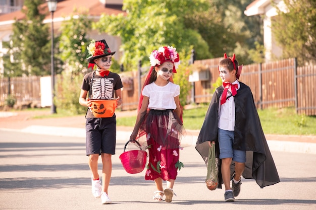 Grupo de crianças simpáticas e fofas em fantasias e pinturas de halloween, batendo papo enquanto se moviam entre casas de campo