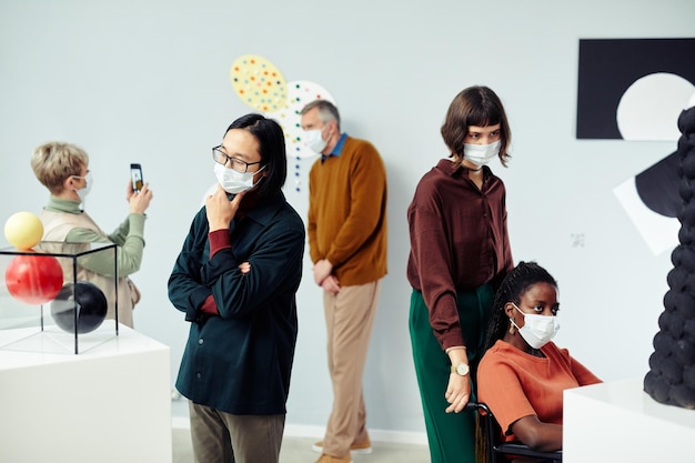 Grupo de diversas pessoas multiétnicas usando máscaras protetoras examinando vários objetos de arte contemporânea em exposição no museu
