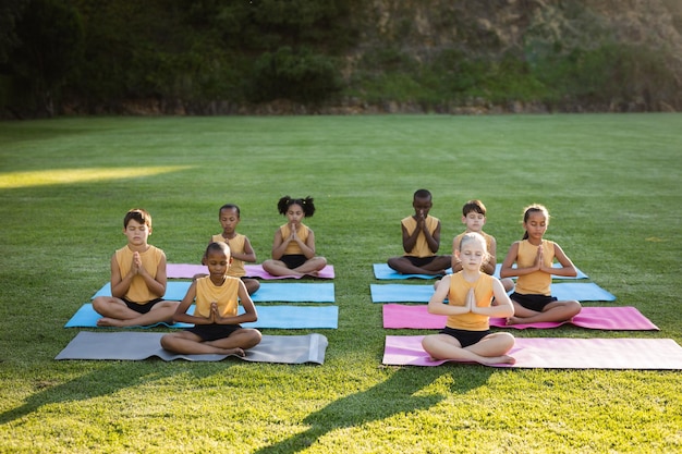 Grupo de diversos alunos praticando ioga e meditando sentado no tapete de ioga no jardim da escola