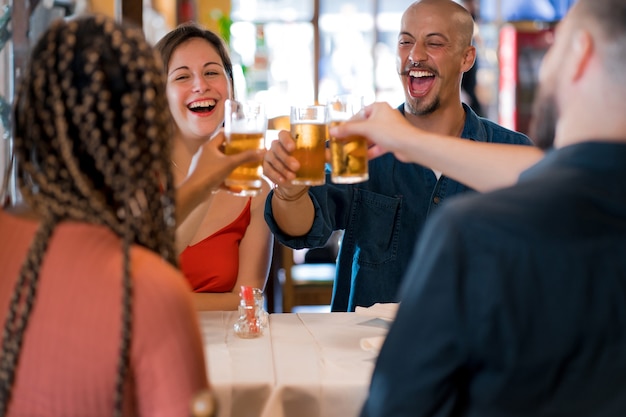 Grupo de diversos amigos brindando com copos de cerveja enquanto desfrutam de uma refeição juntos em um restaurante. Conceito de amigos.