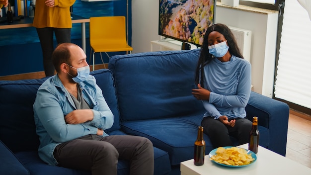 Grupo de diversos amigos reunidos na sala de estar em casa celebrando durante a pandemia global, usando máscara para manter distância social. pessoas multiétnicas tirando máscara de proteção e comendo serpentes