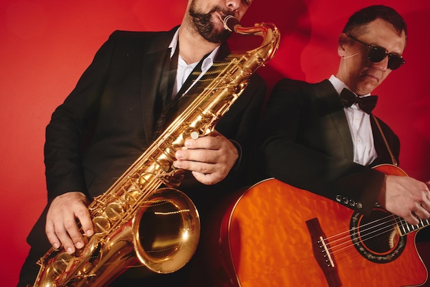 Grupo de dois músicos, banda de jazz masculina, guitarrista e saxofonista em trajes clássicos improvisar em instrumentos musicais em um estúdio em fundo vermelho