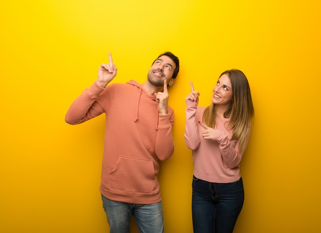 Grupo de duas pessoas em fundo amarelo, apontando com o dedo indicador