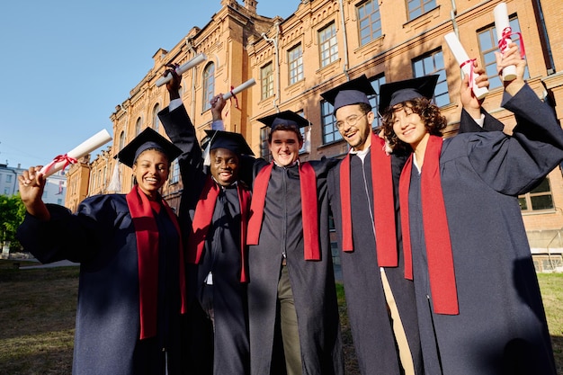 Grupo de estudantes felizes em vestidos com diplomas ao ar livre perto da universidade