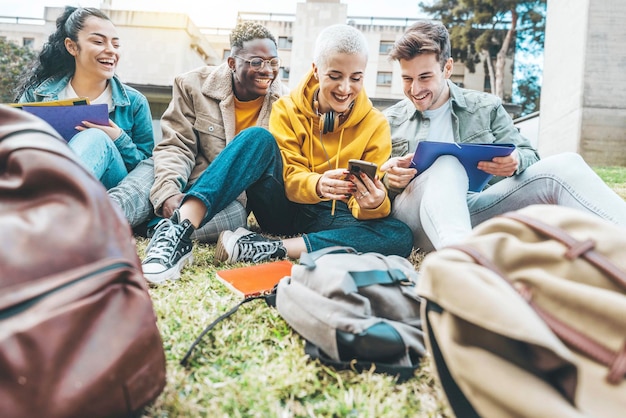 Grupo de estudantes multirraciais se divertindo sentados juntos no campus da faculdade Adolescentes felizes usando telefone celular inteligente do lado de fora Pessoas do milênio rindo ao ar livre Conceito de bolsa de estudos