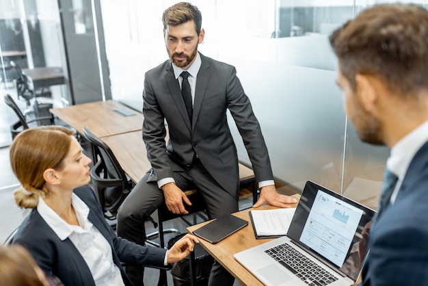 Grupo de executivos estritamente vestidos discutindo gráfico financeiro apresentado no laptop, reunindo-se no escritório moderno com divisórias de vidro