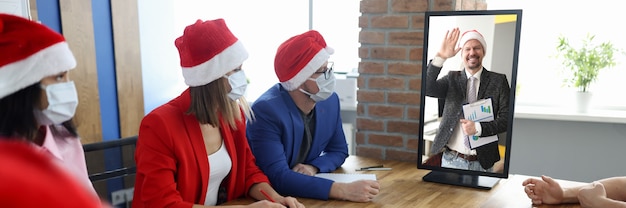 Grupo de homens e mulheres de negócios com máscaras protetoras e chapéus de Papai Noel estão sentados à mesa e