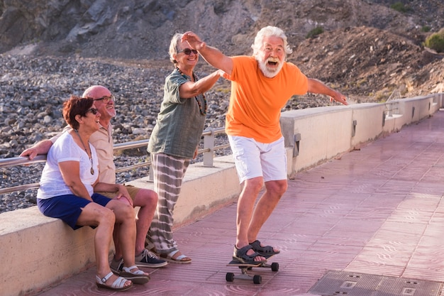 Grupo de idosos e pessoas maduras na praia se divertem olhando o velho andando de skate e rindo com cara de susto - mulher tocando o homem
