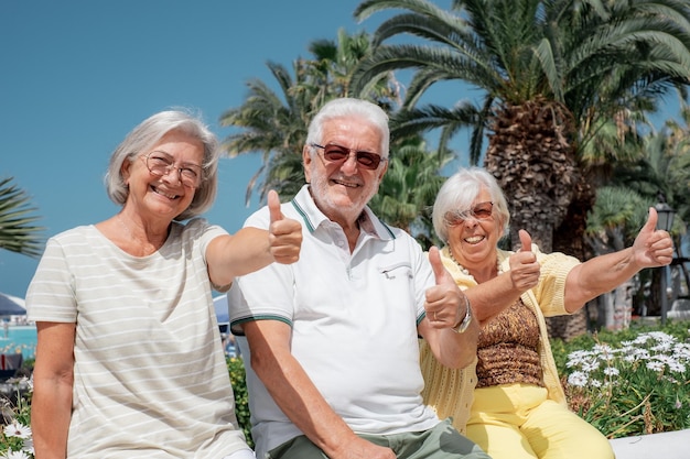 Grupo de idosos felizes com polegares para cima, sentados sob o sol, aproveitando as férias no mar e a liberdade