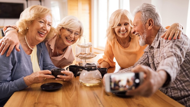 Grupo de idosos felizes usando telefone celular e tomando selfie durante a hora do chá em casa