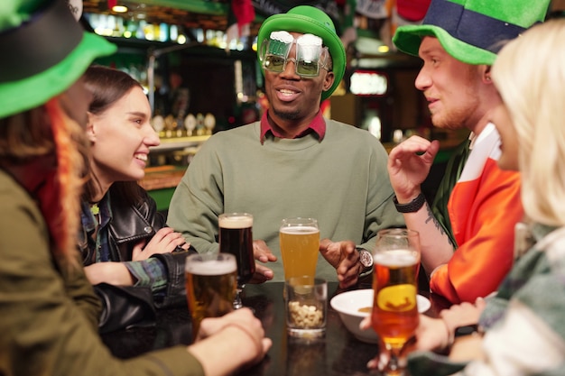 Grupo de jovens amigos interculturais felizes em trajes tradicionais irlandeses tomando cerveja com lanches na frente da câmera enquanto celebra o dia de São Patrício