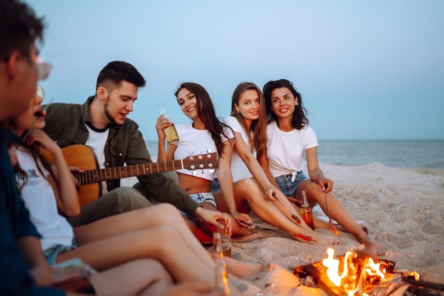 Grupo de jovens amigos sentados na praia e frite salsichas Um homem está tocando violão Hora de acampar