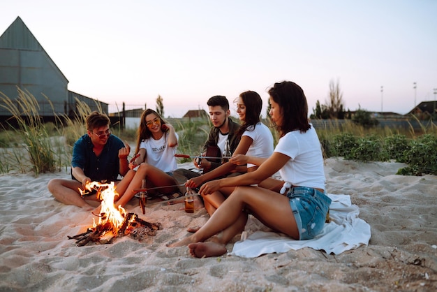 Grupo de jovens amigos sentados na praia e frite salsichas Um homem está tocando violão Hora de acampar