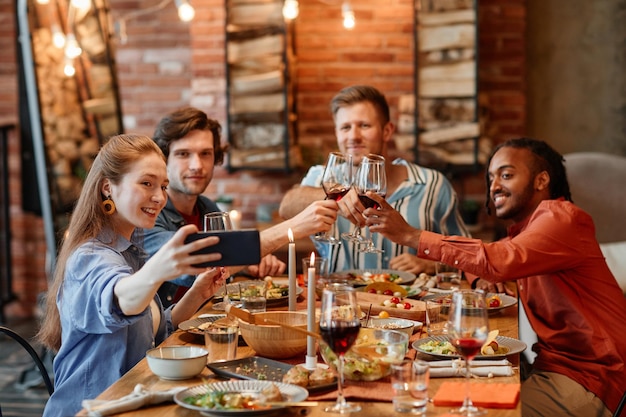 Grupo de jovens amigos tirando foto selfie na mesa durante o jantar e tinindo copos