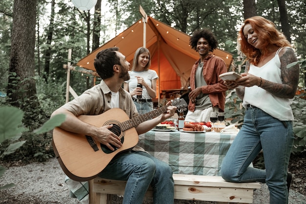 Grupo de jovens amigos viajando em glamping na floresta se divertindo tocando violão e mbira kalimba assando salsichas, sentados à mesa de jantar perto da barraca durante as férias de verão rindo, curtindo