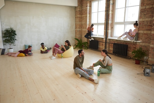 Grupo de jovens dançarinos relaxando após o treinamento esportivo, ouvindo música e usando telefones celulares no estúdio de dança